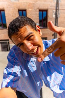 Young man smiling into camera and pointing down in a bright outdoor street selfie, casual striped
