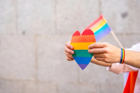 Person's hands holding a rainbow colored heart and a small pride flag, with a rainbow bracelet on