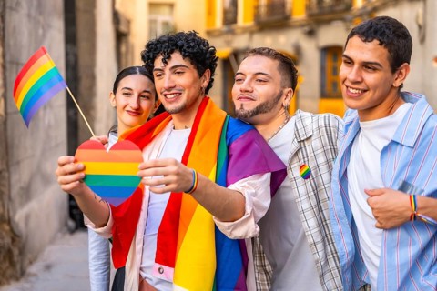 Young friends proudly displaying rainbow heart, flag and shawl at an outdoor pride celebration,