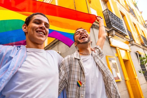 Happy young gay couple holding a rainbow flag and cheering loudly during a pride parade, expressing
