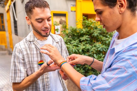 Two men clasp hands and put on rainbow flag bracelets outdoors, celebrating love, pride and