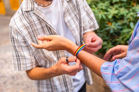 Two hands fastening a colorful rainbow bracelet on a wrist outdoors, symbolizing pride, love and