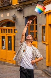 Young man standing on a street, smiling and raising a small rainbow flag, expressing happiness and