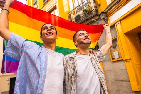 Happy young men joyfully embracing and holding a colorful rainbow flag together, looking up in a