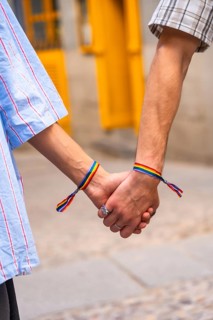Couple's hands holding together, both wrists featuring rainbow bracelets, symbolizing love, pride,