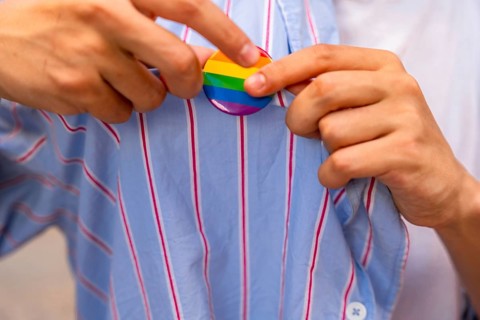 Person's hands carefully pinning a rainbow pride flag button onto a blue and white striped shirt,