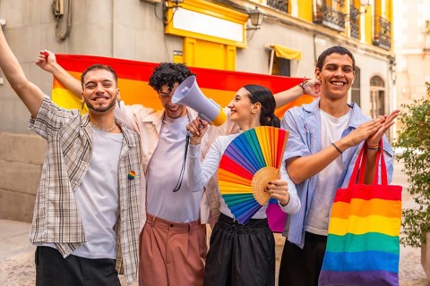 Diverse group of young friends standing together, holding a rainbow flag, fan, and megaphone,