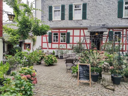 A rustic courtyard with a half-timbered house, surrounded by plants and flowers on cobblestones,