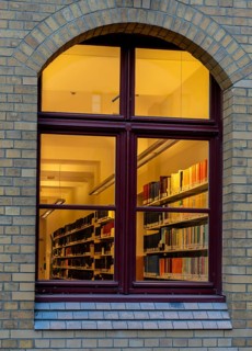 A view through the window of a library, shelves full of books illuminated by a warm, yellow light,