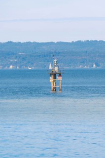 A lighthouse in blue water with a sailing ship, green hills and clear sky in the background,