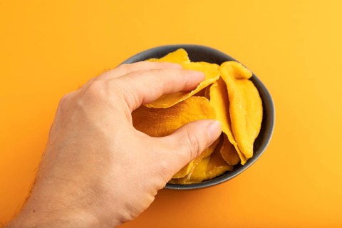 Dried Mango in blue ceramic bowl with hand on orange pastel paper background. Side view, close up.