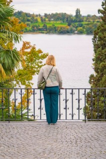 Person standing at the railing of a walkway and looking at a lake surrounded by trees in autumn,