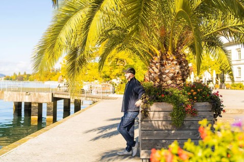 A man leaning against a flower trough on a sunny promenade by the water, Überlingen, Lake
