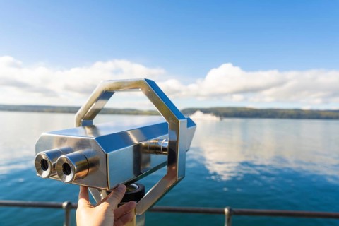 Hand holding binoculars on the lakeshore under a clear sky with clouds, Überlingen, Lake Constance,
