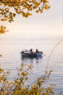 Two people fishing on a small boat in a calm lake surrounded by autumn leaves, Überlingen, Lake