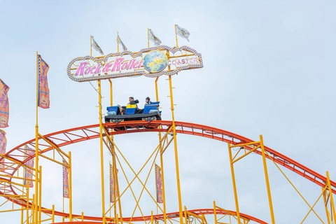 People in a roller coaster car on an orange-coloured track in front of colourful flags, Cannstatter