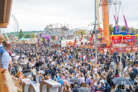 Large crowd at a folk festival with fairground rides under a cloudy sky, Cannstatter Wasen folk