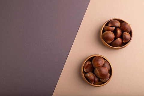 Wooden bowl with raw edible ?hestnuts on brown and gray paper pastel background, top view, flat