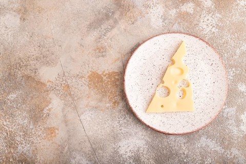 Piece of cheese with holes on ceramic plate on brown concrete background, top view, flat lay, copy