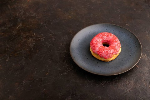 Pink Donut with sprinkles on blue ceramic plate on black concrete background, side view, copy