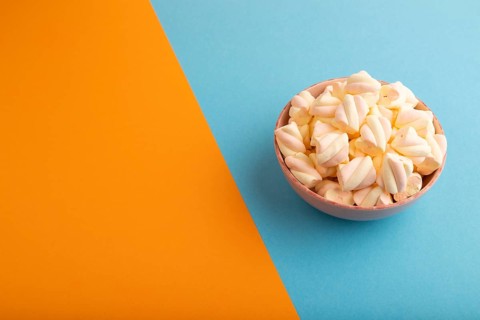 Orange and pink marshmallow in ceramic bowl on blue and orange pastel paper background, side view,
