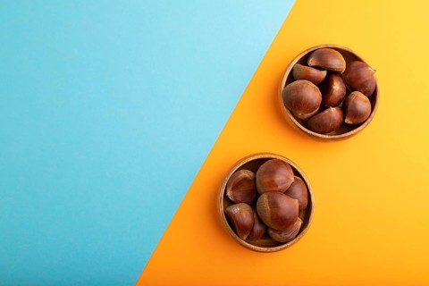 Wooden bowl with raw edible ?hestnuts on blue and orange paper pastel background, top view, flat