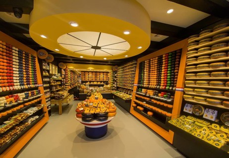 A shop with a variety of cheeses arranged on shelves and tables, illuminated in warm light, Delft,