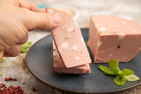Mortadella sausage with hand on plate with pepper and herbs on brown concrete background and linen
