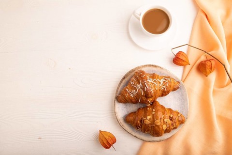 Croissant on blue plate on white wooden background and orange linen textile, cup of coffee, top