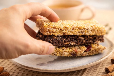 Granola bars with caramel, nuts, flakes in ceramic plate on brown concrete background with hand,