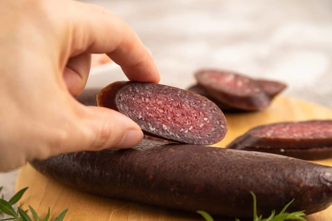 Sujuk sausage with hand on wooden cutting board with pepper and herbs on brown concrete background.