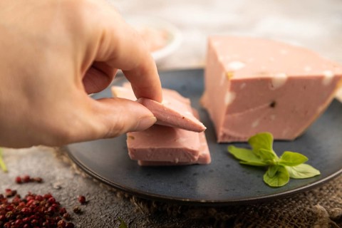 Mortadella sausage with hand on plate with pepper and herbs on brown concrete background and linen