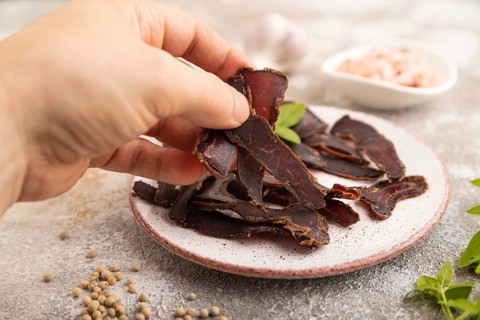 Armenian Basturma dried meat with hand on plate with pepper and herbs on brown concrete background.