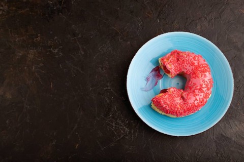Bitten Pink Donut with sprinkles on blue ceramic plate on black concrete background, top view, flat