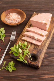 Mortadella sausage on wooden cutting board with pepper and herbs on brown wooden background. Side