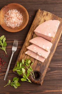 Mortadella sausage on wooden cutting board with pepper and herbs on brown wooden background. Top