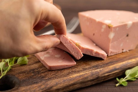 Mortadella sausage with hand on wooden cutting board with pepper and herbs on brown wooden