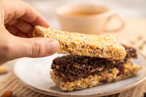 Granola bars with caramel, nuts, flakes in ceramic plate on brown concrete background with hand,