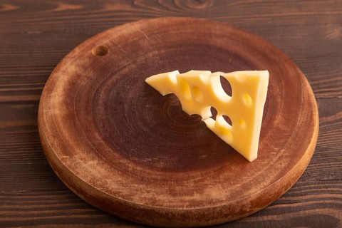 Piece of cheese with holes on cutting board on brown wooden background, side view, close up,