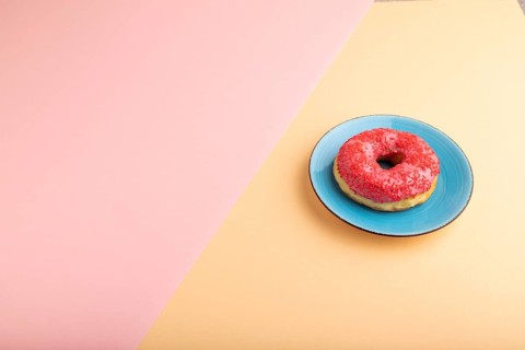 Pink Donut with sprinkles on blue ceramic plate on blue and orange pastel paper background, side