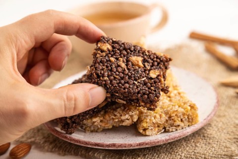 Granola bars with caramel, nuts, flakes in ceramic plate with hand on white wooden background,