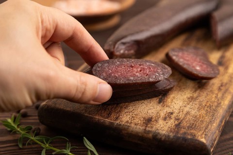 Sujuk sausage with hand on wooden cutting board with pepper and herbs on brown wooden background.