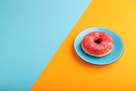 Pink Donut with sprinkles on blue ceramic plate on blue and orange pastel paper background, side