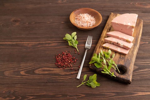 Mortadella sausage on wooden cutting board with pepper and herbs on brown wooden background. Side