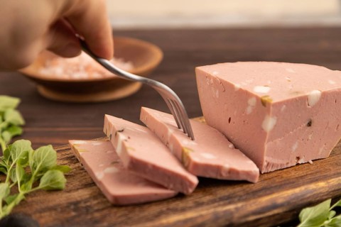 Mortadella sausage with hand on wooden cutting board with pepper and herbs on brown wooden