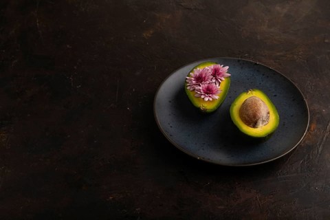 Two Avocado halves with pit and Chrysanthemums on blue plate on black concrete background, side