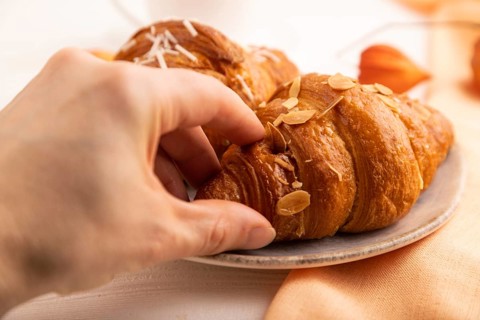 Croissant on blue plate with hand on white wooden background and orange linen textile, side view,