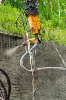 Nahaufnahme einer Maschine, die Beton auf eine Struktur sprüht, umgeben von grüner Vegetation, Bau