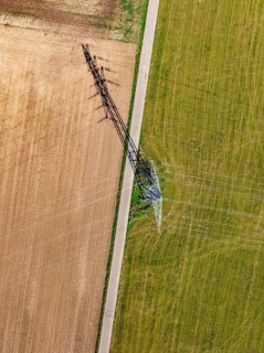 Strommast thront zwischen Acker und Wiese, mit einem langen Schatten der sich über das Feld