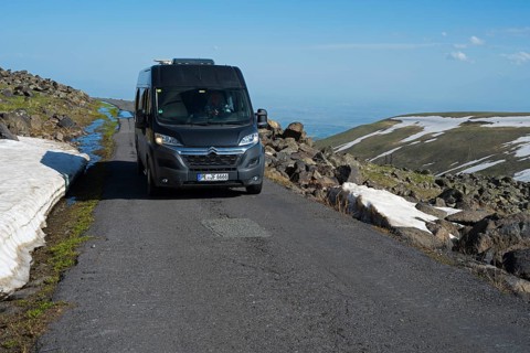 Van fährt auf asphaltiertem Weg neben Felsen und Schneefeldern mit klarem Himmel, das Tal im